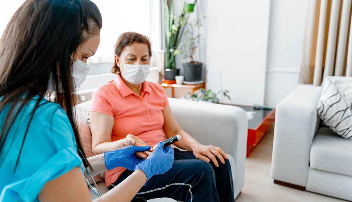 A home health aide wearing a face mask and gloves testing a woman's blood sugar at home.