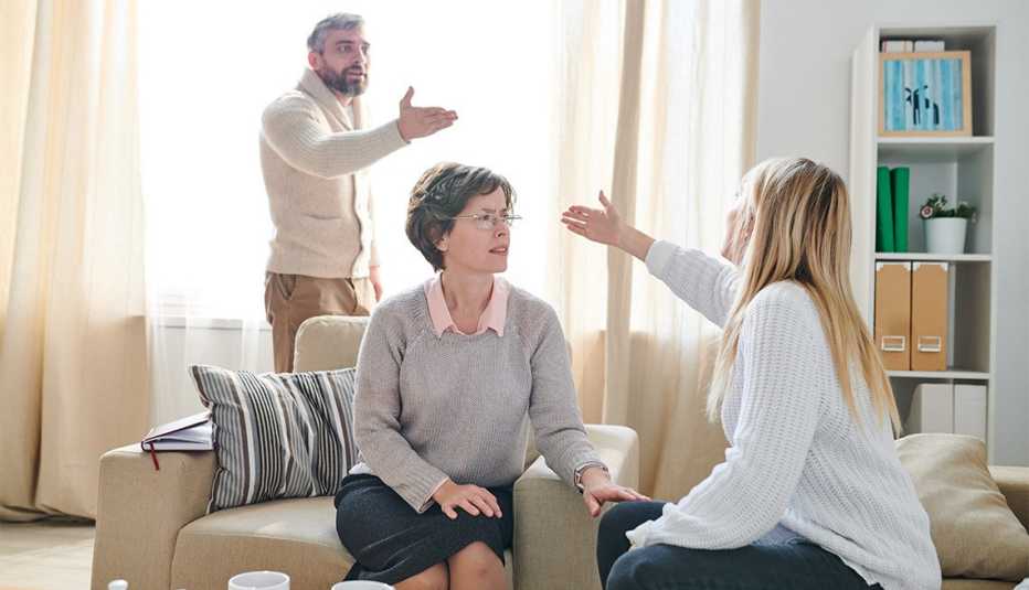 Family psychologist in glasses sitting in armchair and listening to quarrel of couple while asking young lady to stop conflict, emotional couple gesturing hands and blaming each other for their problems