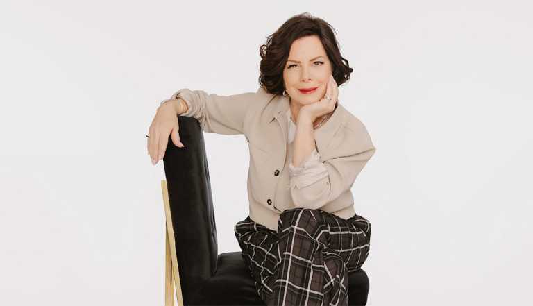 Marcia Gay Harden sitting on chair with right arm leaning on top of chair and face resting on left hand; white background