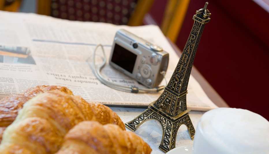 overhead shot of a cafe table filled with a newspaper, camera, small metal eiffel tower souvenir foamy coffee drink and croissants
