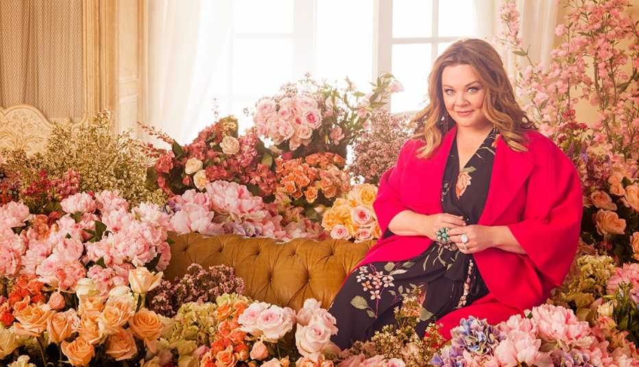 Melissa McCarthy sitting on a couch surrounded by flowers