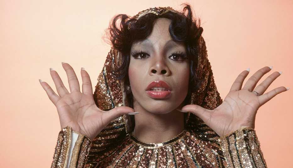Headshot of Donna Summer, US singer-songwriter, posing in a studio portrait, with her hands raised, open-palmed, either side of her head, wearing gold-sequinned cuffs and collar, 1976.