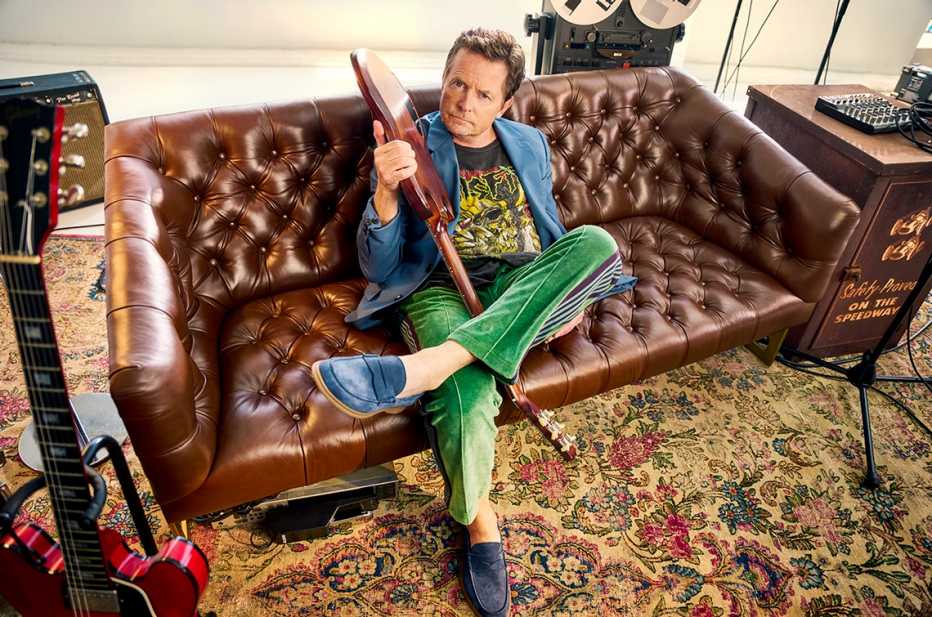 michael j fox sitting on a couch wearing trendy clothes holding a guitar
