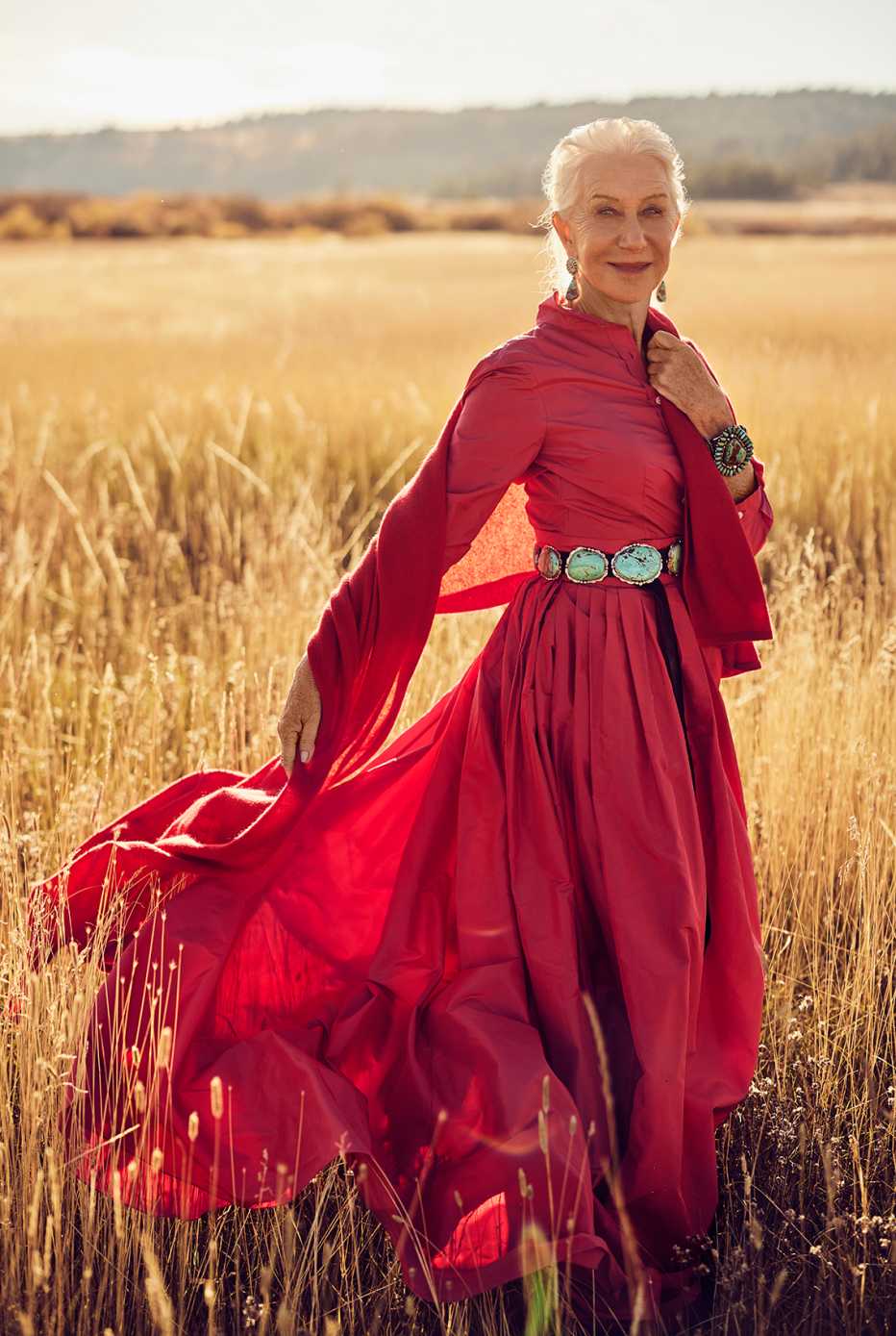 Helen Mirren in a red dress with Western accent pieces posing in a sunlit field