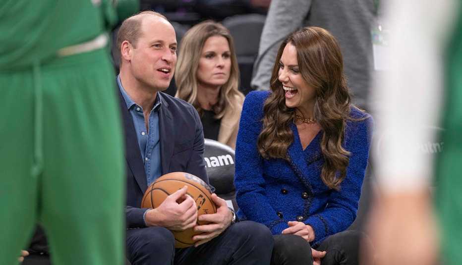 Prince William, Prince of Wales, holds a basketball in his lap while Catherine, Princess of Wales, laughs alongside him at a Boston Celtics game