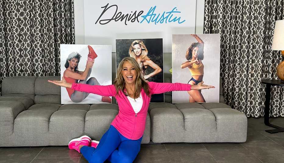 denise austin lifts her arms out laterally while sitting on the floor with three portraits of her younger self placed behind her on a couch and a denise austin logo above