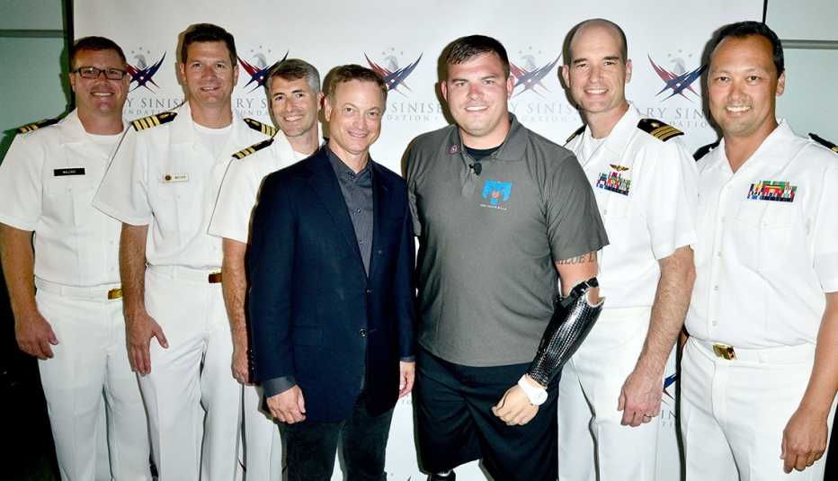 gary sinise, wearing a dark blue suit, and veteran travis mills, wearing a gray polo and dark shorts, pose with five military members in dress whites