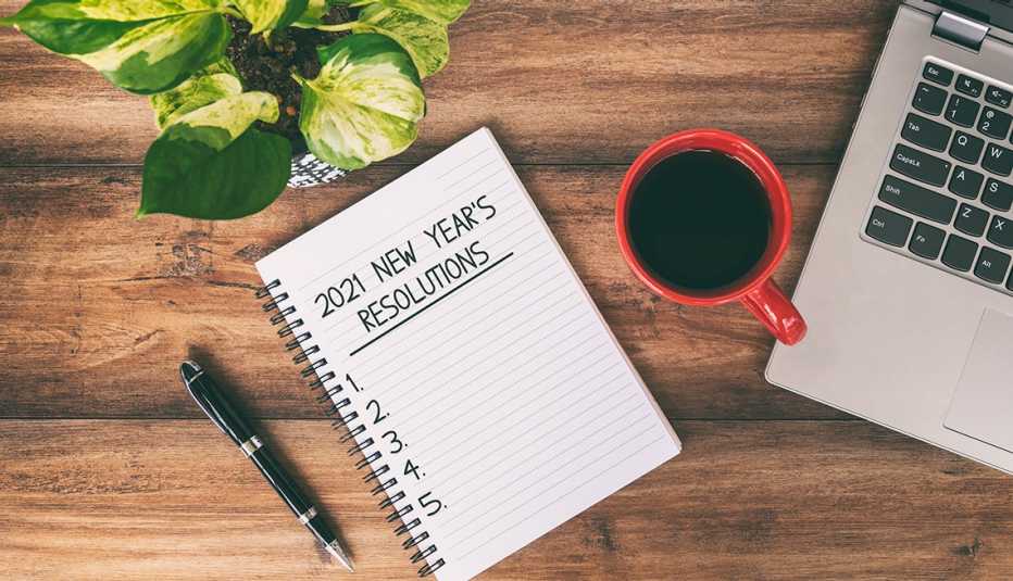 A notepad with 2021 New Year's Resolutions written on a page with a laptop and a cup of coffee nearby on top of a wooden desk