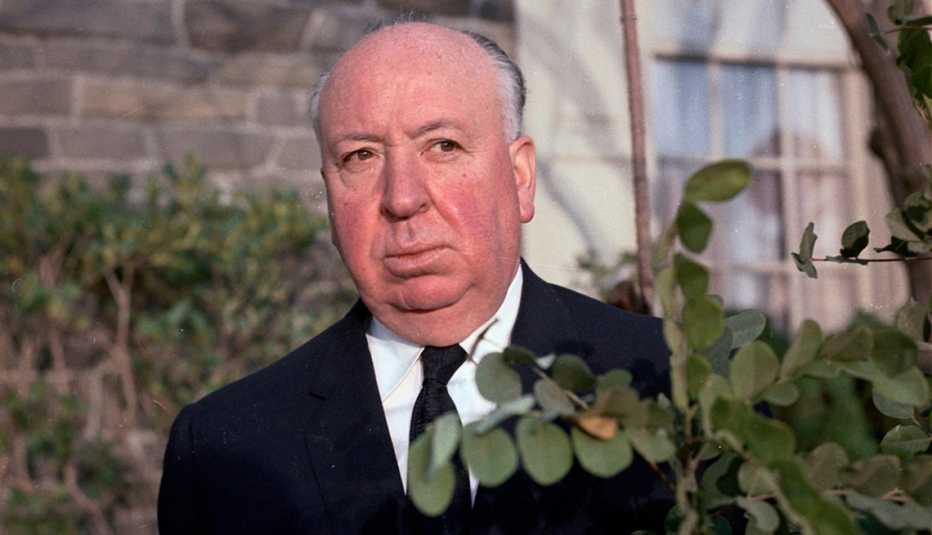 Director Alfred Hitchcock standing by a bush