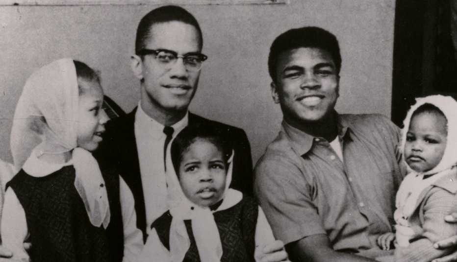 Malcolm X and Muhammad Ali sitting next to each other while holding their daughters in their laps
