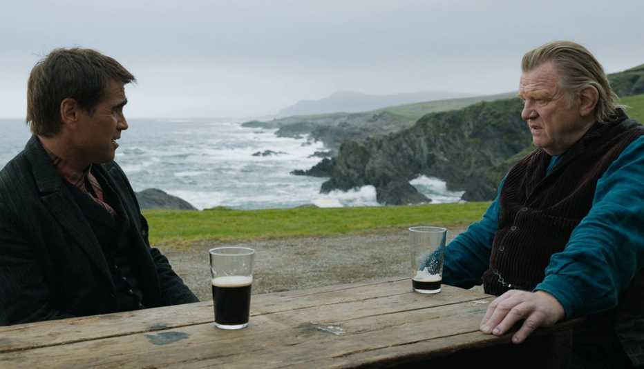 Colin Farrell and Brendan Gleeson sitting at a table with glasses of beer while overlooking the water in the film The Banshees of Inisherin