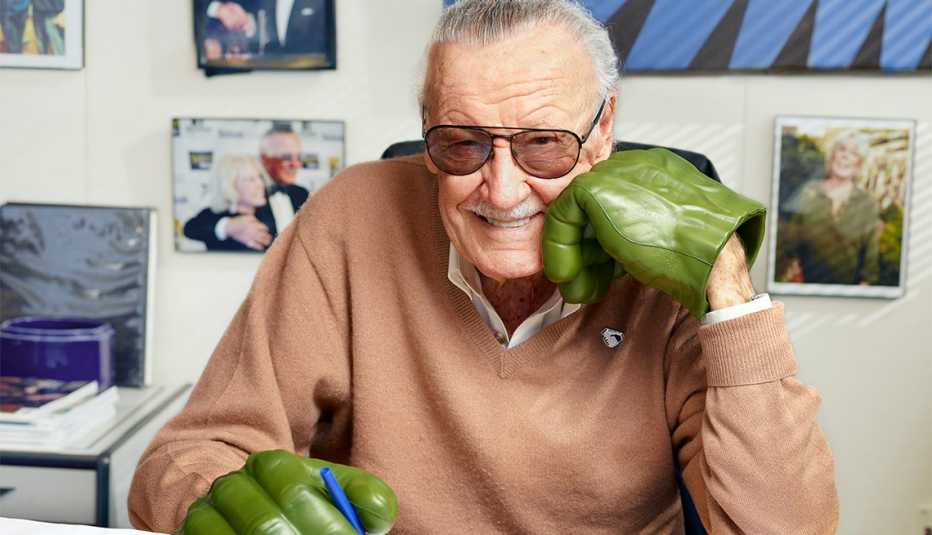 stan lee smiles at his desk holding a pen wearing green hulk gloves 