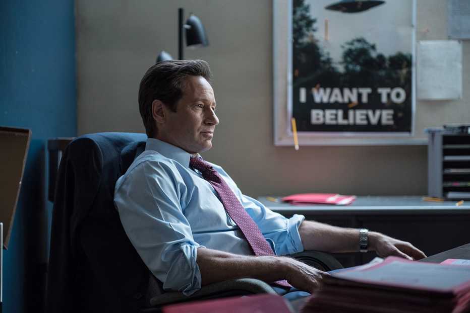 David Duchovny sitting in a desk chair with a I Want to Believe poster nearby him in the background