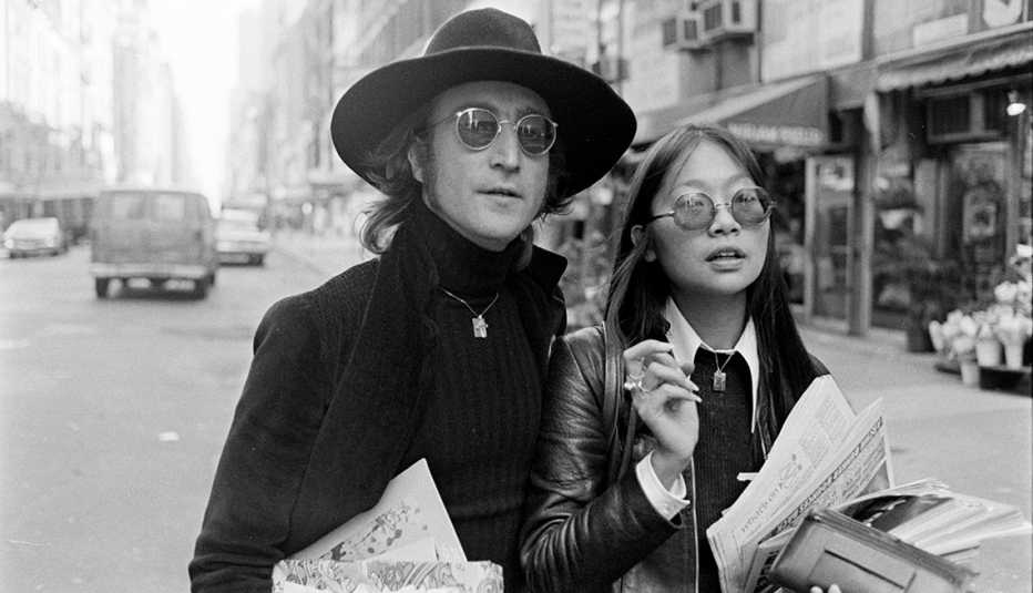 john lennon and may pang walk down a new york city street, both wearing sunglasses and carrying papers
