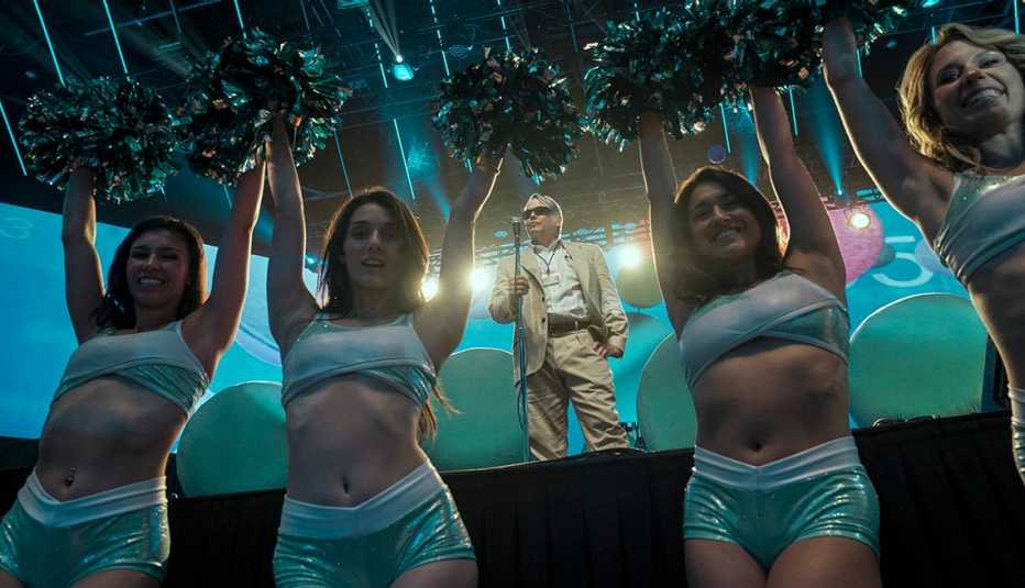 Matthew Broderick stands on a stage in front of a microphone as cheerleaders stand in front of the stage in the Netflix series Painkiller