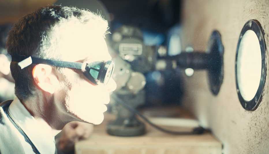 cillian murphy wearing googles while looking into a hole emitting a bright light in the film oppenheimer