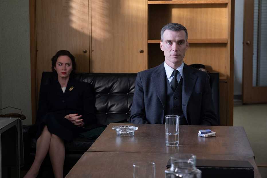 emily blunt sitting on a couch behind cillian murphy as he is sitting in a chair at a desk in the film oppenheimer