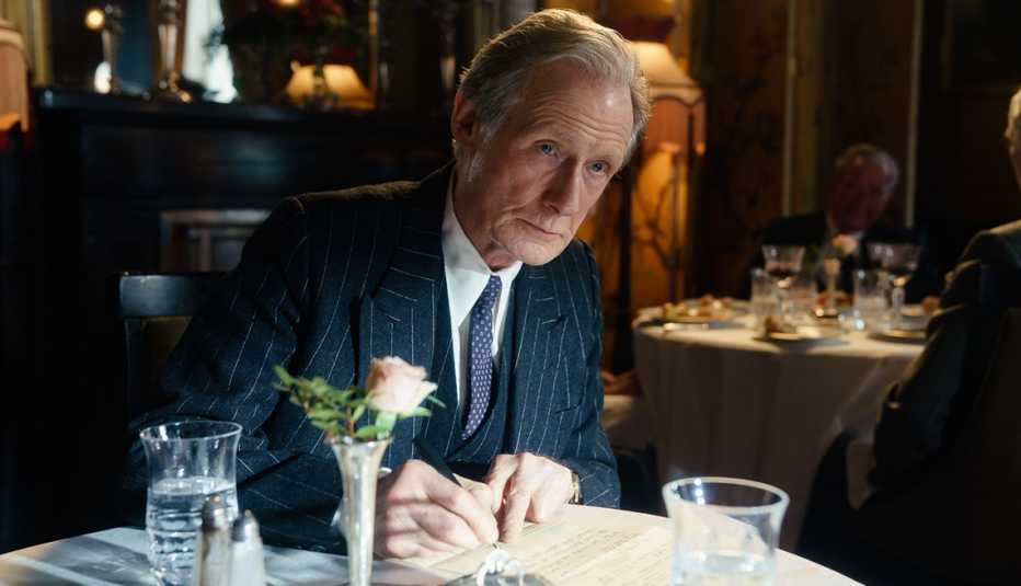 Bill Nighy sitting at a restaurant table writing with a pen in the film Living