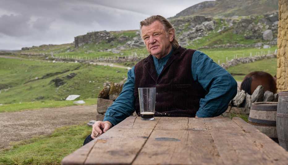 Brendan Gleeson sitting at a wooden table outdoors with a glass of beer in front of him in the film The Banshees of Inisherin