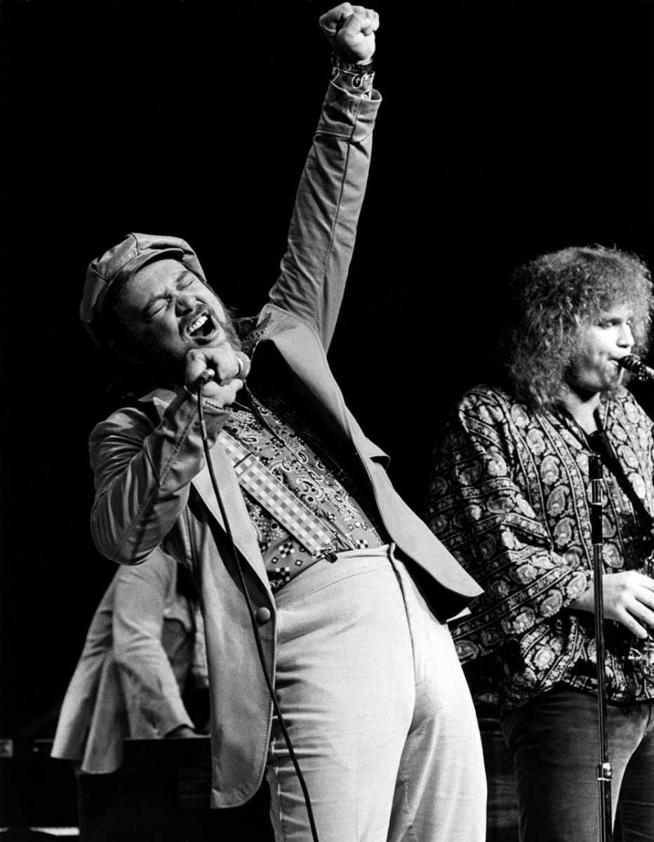 david clayton thomas singing into a microphone with his left fist in the air while performing onstage with his band blood sweat and tears