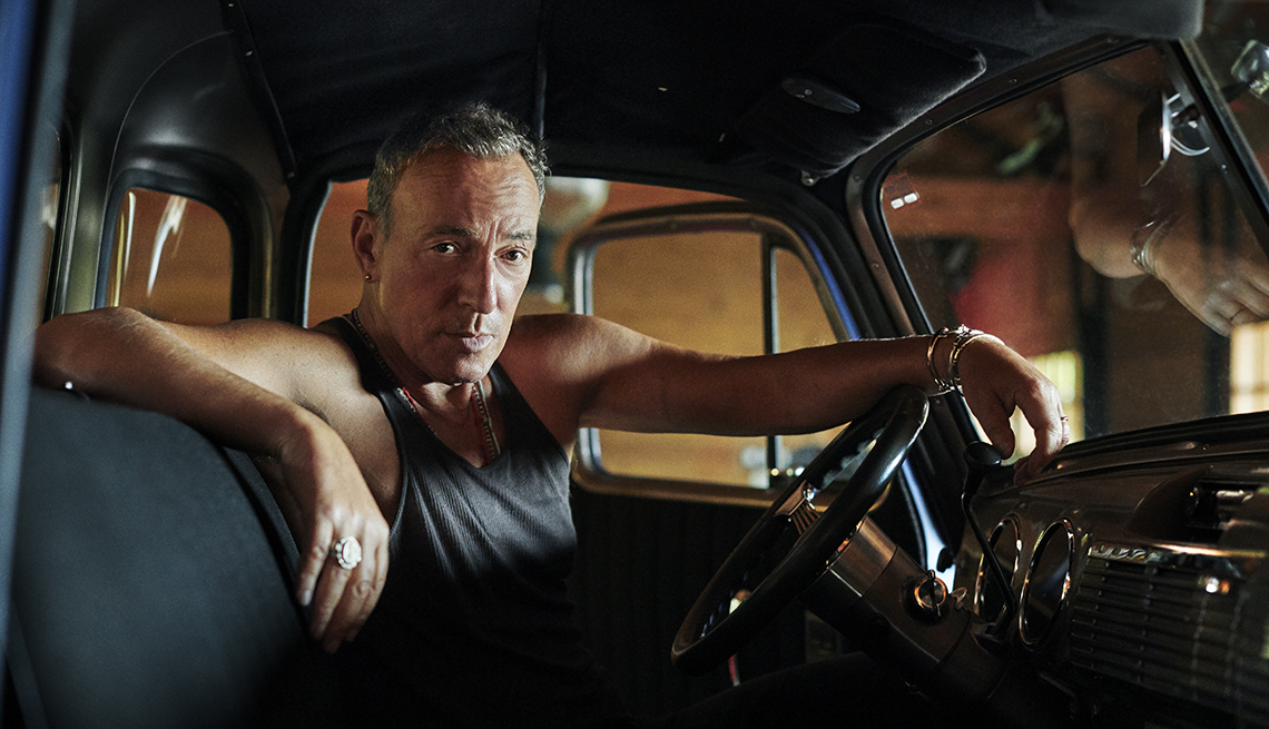 bruce springsteen poses in the drivers seat of one of his antique cars