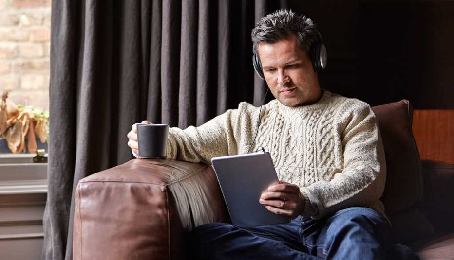 A man sitting on a sofa listening to audio on headphones while holding a coffee mug
