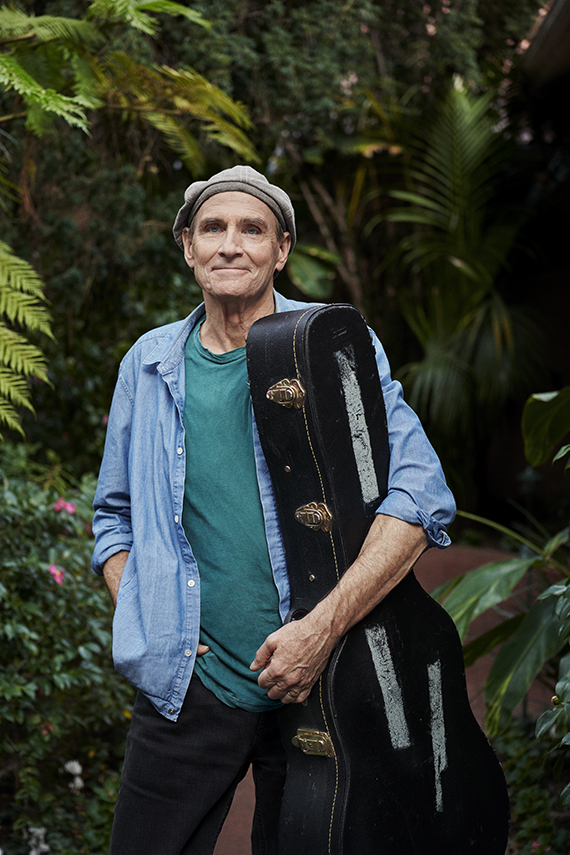 musician james taylor holding his guitar case and smiling in front of a garden backdrop