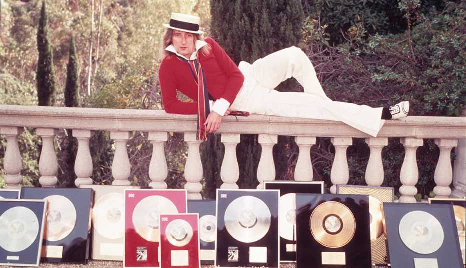 Rod Stewart lying on an outdoor railing with framed gold and silver record displayed on the ground