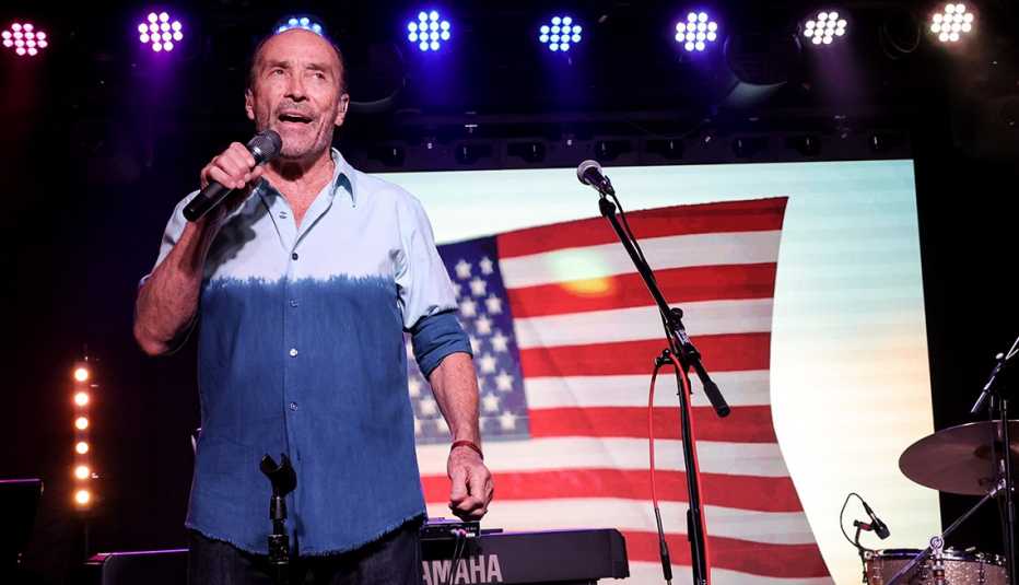 lee greenwood holds a microphone and sings in front of an american flag