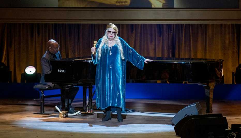Joni Mitchell holding a microphone in front of a piano onstage during her performance at the 2023 Library of Congress Gershwin Prize for American Song ceremony