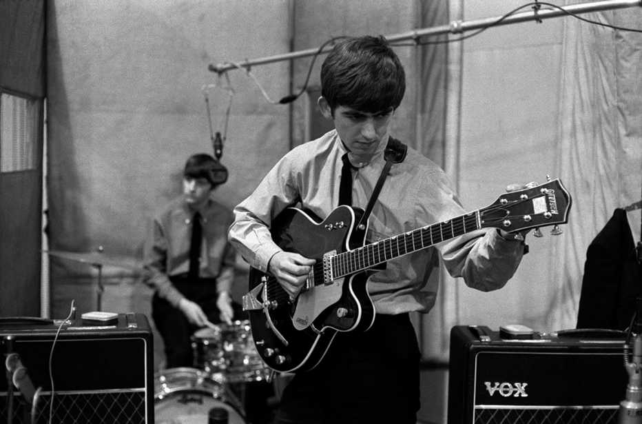 George Harrison tuning his guitar with Ringo Starr seated at his drum set in the background while recording She Loves You at Abbey Road Studios in London