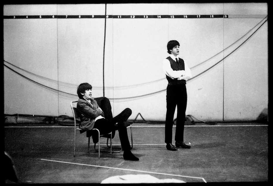 George Harrison sitting in a chair while Paul McCartney stands next to him with his arms crossed at a Beatles rehearsal for a TV special at Wembley Studios in April 1964