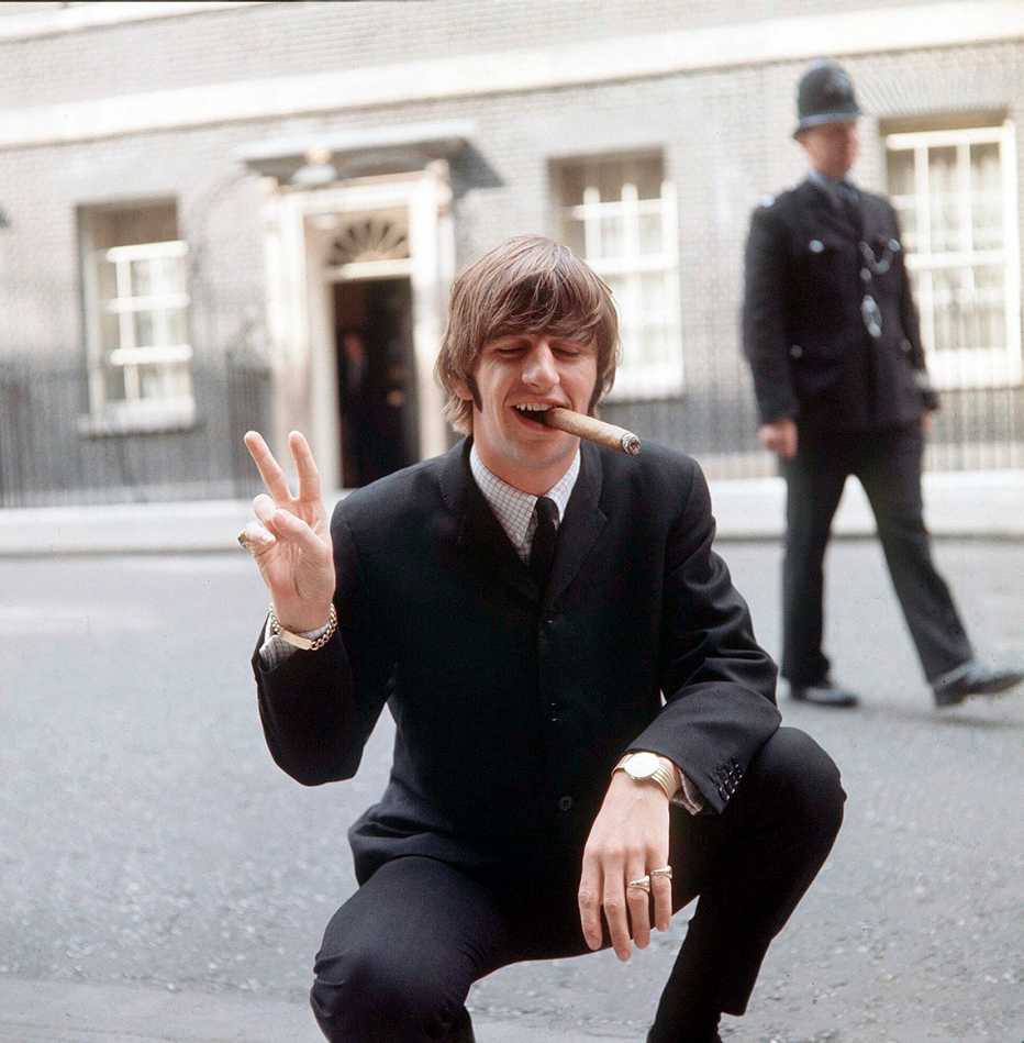 Ringo Starr makes a peace sign while squatted outside on a street with a cigar in his mouth