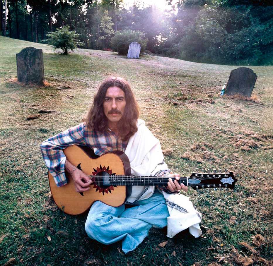 George Harrison sitting on the ground with his acoustic guitar nearby gravestones on the grounds of his home in Friar Park