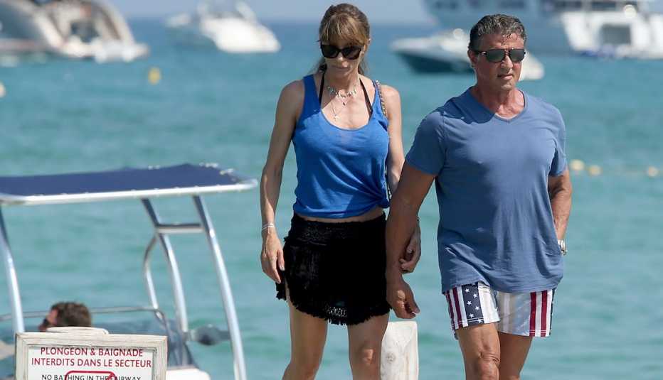 Sylvester Stallone and Jennifer Flavin beachside - he's in patriotic stars and stripes shorts, a gray blue t-shirt and black kicks, she's in a blue tank, black ruffled mini wrap over a halter bikini...no envy please.