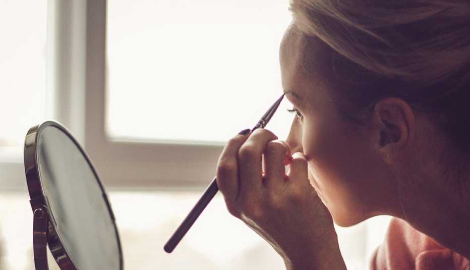 A woman using a brow pencil in front of a small mirror