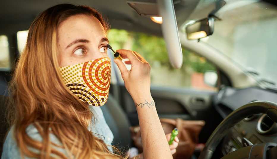 A woman wearing a face mask is applying makeup sitting in the driver seat of her car