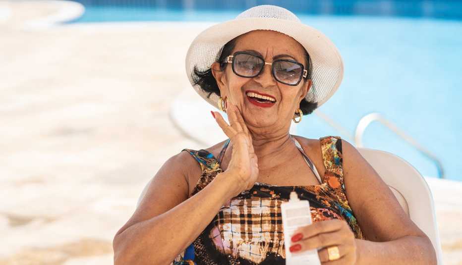 smiling woman sitting in shade at pool wearing sunglasses and hat and applying sunscreen