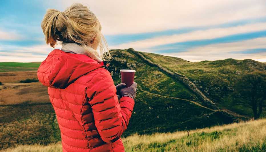 a woman in a red winter coat holds coffee while looking at the countryside