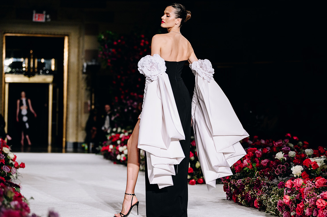 A model on the runway turned with her back to the audience showing her wearing a black dress with two white bows on her arms at Christian Siriano RTW Fall 2023 fashion show