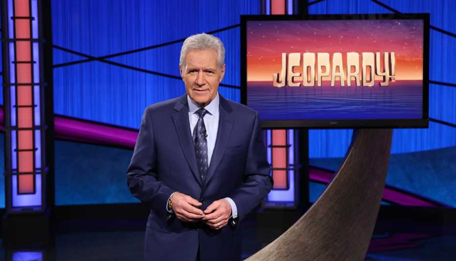 Alex Trebek in a studio standing in front of a Jeopardy monitor