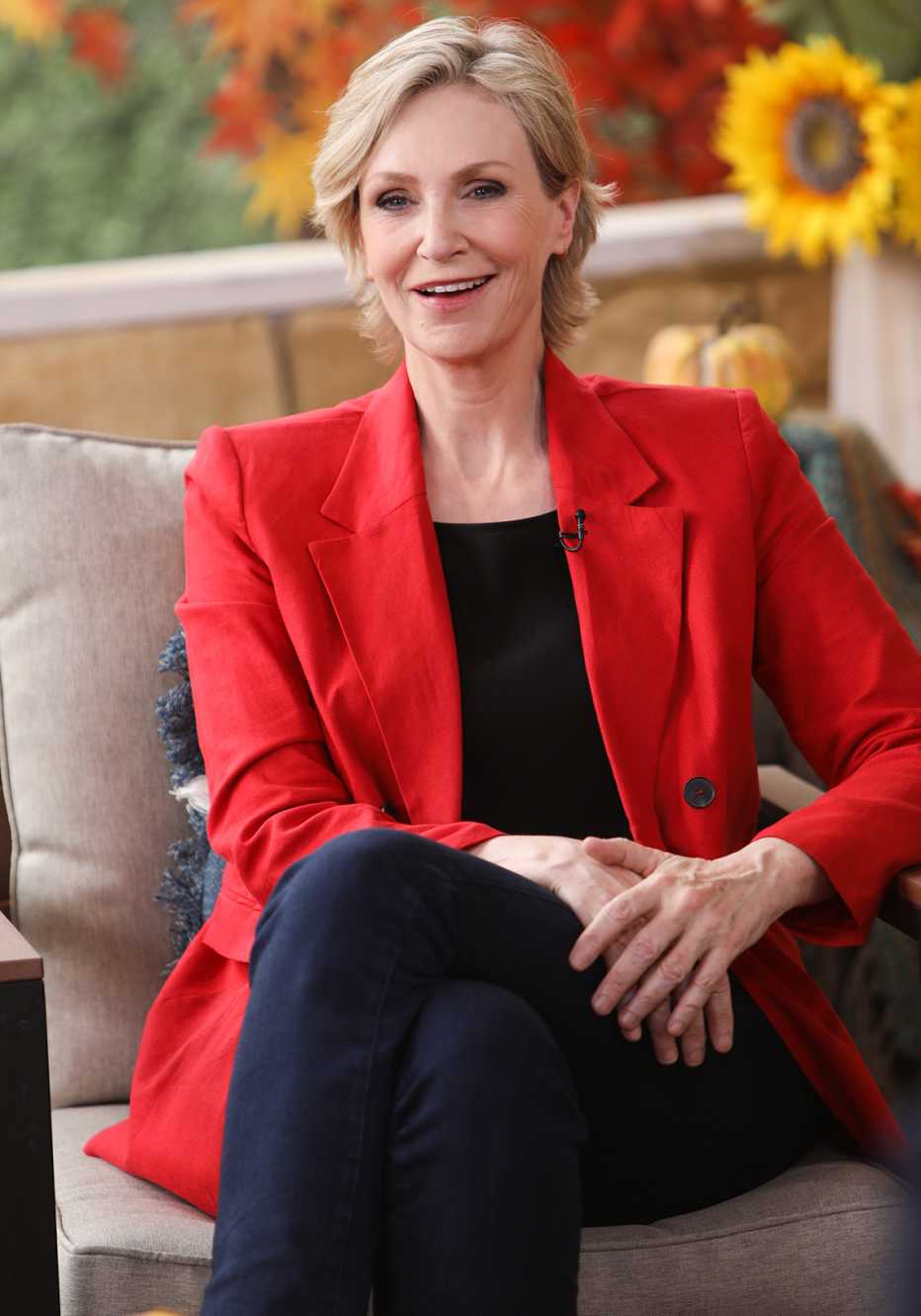 Actress Jane Lynch sitting in a chair in a fall-themed background setting