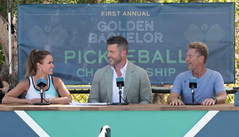 trista sutter jesse palmer and gerry turner sit in front of microphones at a table with a sign that says first annual golden bachelor pickleball championship behind them