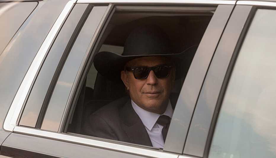 Kevin Costner sitting inside a vehicle wearing a sunglasses, a suit and sunglasses in Yellowstone