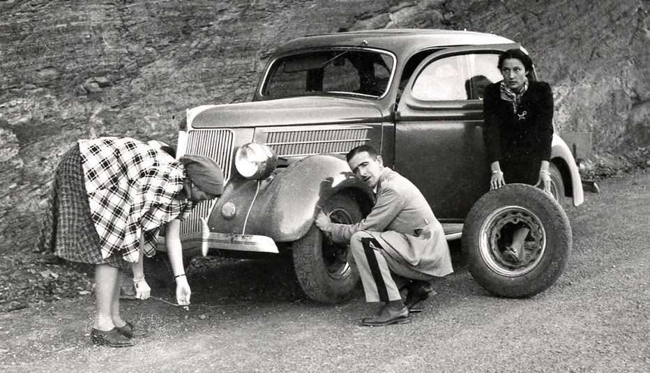 two women and a man change a tire on a road side in a black and white photo