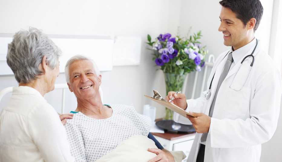 An old couple, the man in a hospital bed, speaking with a smiling doctor.