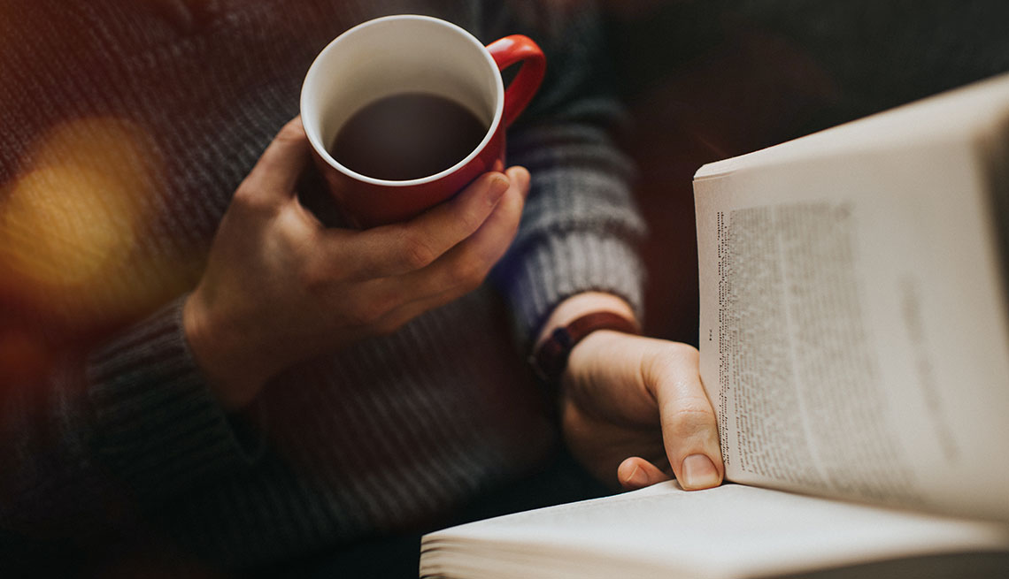 Male reading a book and drinking a hot drink from a red mug.