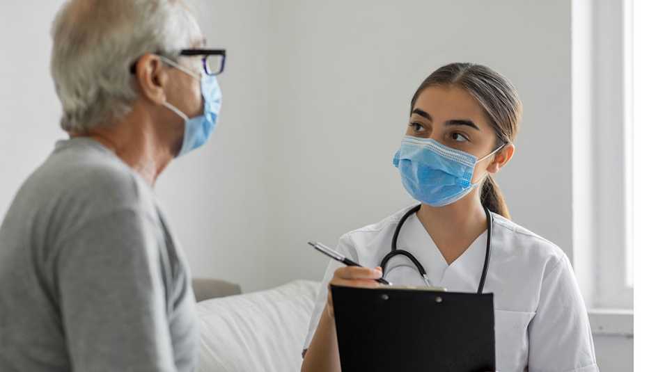 masked older patient speaks with a masked health provider who is taking notes on a clipboard