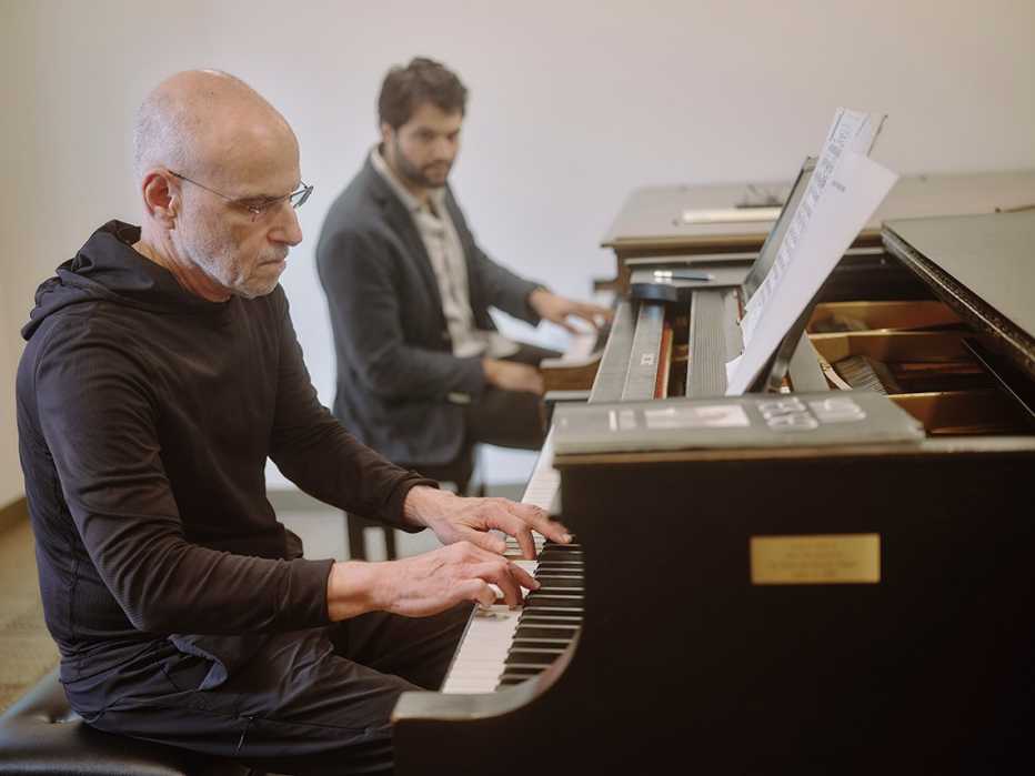 jeffrey galvin plays piano in the foreground while his piano teacher jason solounis plays another piano in the background and looks over at jeffreys playing