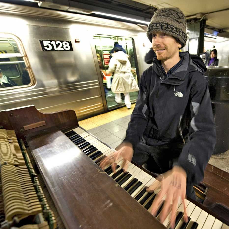 colin huggins plays a piano on the new york subway platform with a train in the background